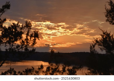 A golden sunset illuminates the sky with warm hues of orange and yellow, casting a soft glow over a calm lake. Silhouetted tree branches frame the peaceful scene, adding depth and tranquility. - Powered by Shutterstock