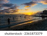 Golden sunset on horizon over Waikiki beach in Oahu, Hawaii, with silhouette of unidentifiable visitors swimming and on the beach.