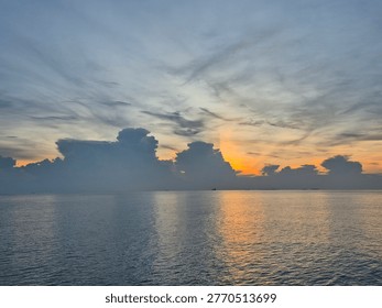 Golden sunset peaking through dramatic clouds over a calm ocean horizon with subtle ship silhouettes.