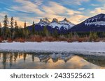 Golden winter sunset at The Three Sisters, a trio of peaks near Canmore, Alberta, Canada, in the Canadian Rockies, with alpenglow on the mountains reflecting off melted snow and ice in the foreground.