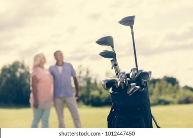 Golf Bag Standing On A Grass Field, Older Couple In The Blurred Background. Active Leisure Time.