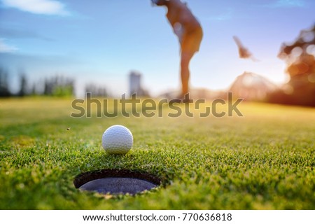 Golf ball approach to the hole on the green, putting by woman golf player in background 