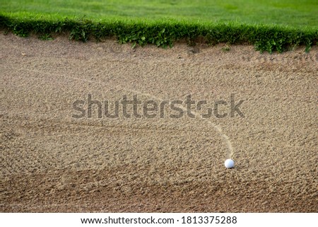 Golf ball lying in a sand bunker with a curved trail visible to show the path it has rolled.
The edge of the bunker with green grass in the background is shown.