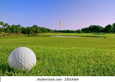 Golf Ball On Bunker Above Flag And Green On Tropical Course In Morning