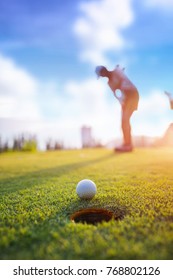 Golf Ball Putting By Woman Golf Player In Background, Golf Ball Spining To The Hold On The Green Of Golf Course With Early Light Of Sunset