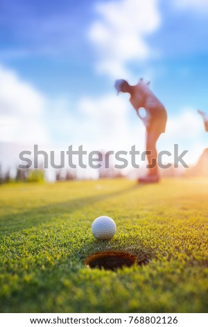 Golf ball putting by woman golf player in background, golf ball spining to the hold on the green of golf course with early light of sunset