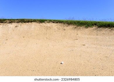 Golf ball in a sand trap, looking up at the top of the bunker and blue sky, recreation and challenge on a sunny summer day
					