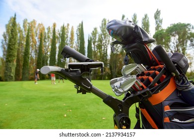 Golf Cart With Bag And Golf Clubs In The Fairway Of A Golf Course, Parked At The Edge Of The Green. In The Background The Players Are Preparing To Pocket The Ball