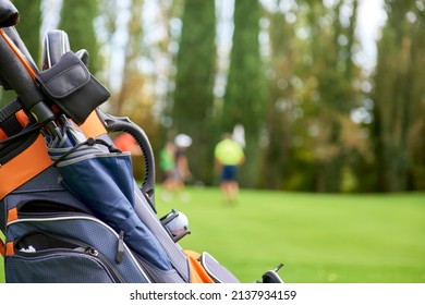 Golf Cart With Bag And Golf Clubs In The Fairway Of A Golf Course, Parked At The Edge Of The Green. In The Background The Players Are Preparing To Pocket The Ball