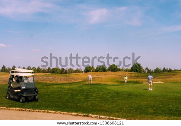 Golf carts parked at the side of the road, golfers\
play golf on a hilly\
field.