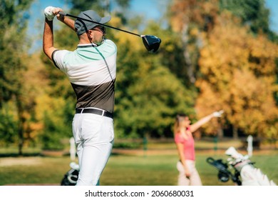 Golf Couples. Young Man Teeing Off, His Partner Following The Ball