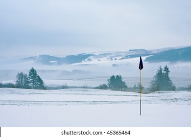 A golf course in Scotland on a snowy winter morning in December. View from a green with a blue flag, with trees, mist and mountains in the background.