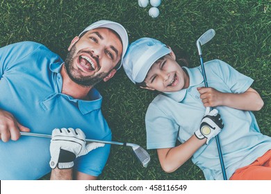 Golf is fun! Top view of cheerful little boy and his father holding golf clubs and smiling while lying on the green grass
