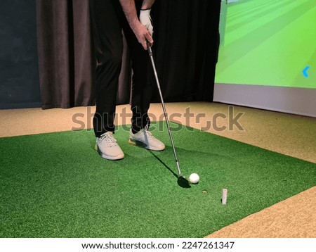 Golf player playing golf indoors on golf simulator closeup. Driving range with screen for golf and golfer legs