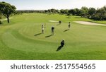 Golf players participating in a game on a well-maintained golf course under a sunny sky. A golfer takes their shot from the green while a player holds a flagstick.