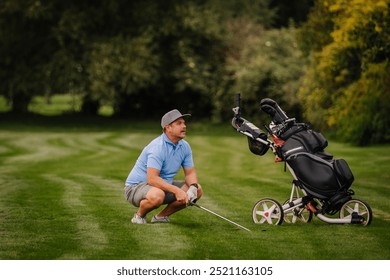 A golfer crouches on the fairway, analyzing his shot while holding a club. A golf cart with bags stands nearby on the green course. - Powered by Shutterstock