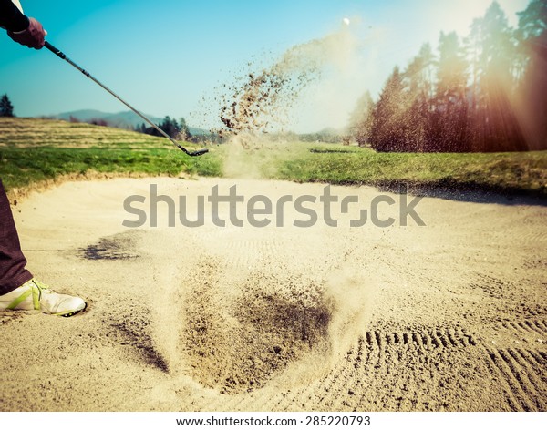 Golfer hitting out of a sand trap. The golf\
course is on the sand. Sand making splashes. Sun and sunshine in\
the background