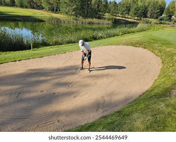 Golfer practicing a bunker shot on a sunny day at a scenic golf course surrounded by trees and a serene pond - Powered by Shutterstock