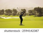 A golfer skillfully swinging a club, aiming at the ball near sand bunker on a bright, sunny day. The lush green setting enhances the serene golf course atmosphere.