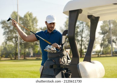Golfer taking putter on golf cart before playing golf on green lawn at warm sunny day. Concept of entertainment, recreation, leisure and hobby outdoors. Young european man in cap