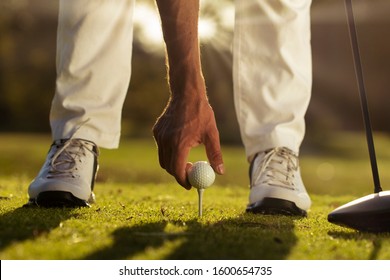 Golfers hand placing golf ball onto tee close up, as he prepares to tee off with a one wood driver, Blurred white golf shoes and white golf pants are also back lit by a sun flare.  - Powered by Shutterstock