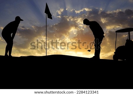 Golfers' hit golf ball toward the hole at sunset silhouetted. Golden morning sky in winter, misty high mountain background.                                