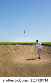 Golfing Middle Aged Man, Hitting Golf Ball Out Of The Bunker. Shot From Inside Bunker With Clear Blue Sky In Background. Copy Space.