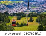 Gondolas ascending to Mount Pilatus, with the city of Kriens near Lucerne in the background, Switzerland, surrounded by lush green fields and forested areas.