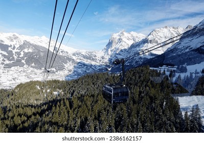 Gondolas of Eiger Express Cableway flying over the forest, with a view of Grindelwald village in the snowy valley and Wetterhorn mountain under blue sky in background, in Berner Oberland, Switzerland