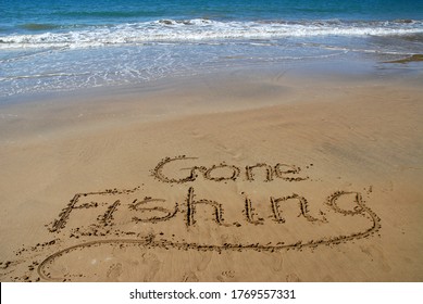Gone fishing written in sand on the beach with surf and blue sea beyond.
