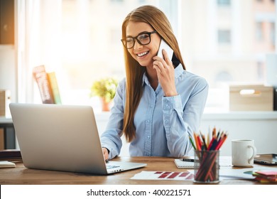 Good business talk. Cheerful young beautiful woman in glasses talking on mobile phone and using laptop with smile while sitting at her working place