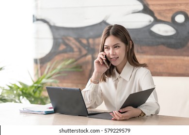 Good business talk. Cheerful young asian girl beautiful asian woman talking on mobile phone and using laptop with smile while sitting at her working place