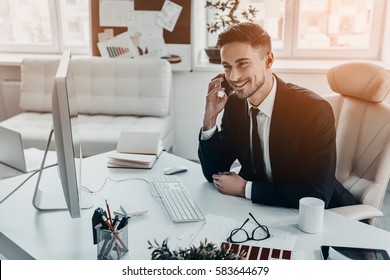 Good Business Talk. Handsome Young Man In Formalwear Talking On The Phone And Smiling While Sitting At The Office Desk 