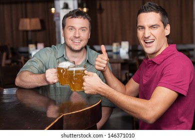 Good Day To Meet Friend. Two Happy Friends Holding The Mugs With Beer And Gesturing On Camera