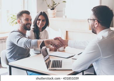 Good deal! Cheerful young man bonding to his wife while shaking hand to man sitting in front of him at the desk 