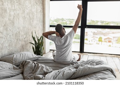 Good Early Morning Concept. Rear Back View Of African American Male Waking Up Sitting On Bed In Bedroom At Home. Millennial Man Stretching Back And Arms, Looking At Window, Free Copy Space - Powered by Shutterstock