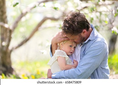 A Good Father Is Holding And Comforting His Sad And Pouting Baby Daughter Outside On A Spring Day.