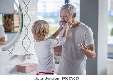 Good Fun Morning. Surprised Cheerful Gray-haired Dad And Child Smearing His Face With Soap Suds Standing Near Mirror