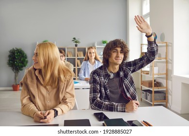 Good High School Or College Student In Casual Wear Asking Or Answering Question In A Lesson. Active Inquisitive Young Man Or Teen Boy With Curly Dark Brown Hair Raising Hand Sitting At Classroom Desk