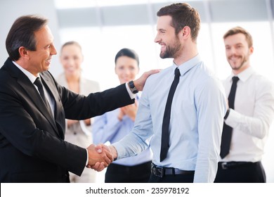 Good job! Two cheerful business men shaking hands while their colleagues applauding and smiling in the background