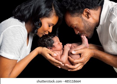 A good looking African American couple cradles their newborn daughter between them in their hands as they both lean in and kiss her.