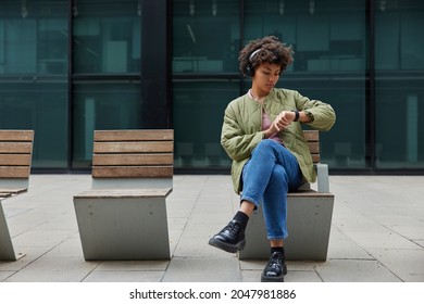 Good looking Afro American woman listens podcast online in wireless headphones enjoys music from playlist waits for someone and looks at smartwatch wears stylish clothes. People and lifestyle concept