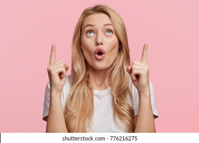 Good looking blonde female with pure healthy skin looks in amazement as indicates at something upwards, isolated over pink background. Pretty young woman sees amazing thing up, gestures indoor
