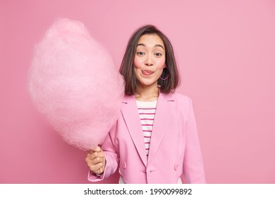 Good looking brunette Asian woman licks lips as holds appetizing sweet candy floss dressed in fashionable formal outfit isolated over pink background. Teenage girl with delicious cotton candy - Shutterstock ID 1990099892