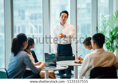 A good looking, confident and fit Asian Chinese man chairs a meeting with his team during the day in the office. He is professionally dressed in a shirt and pants and is gesturing as he speaks. 