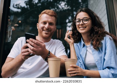 Good looking couple in love smiling at camera coffee meeting in street cafe, portrait of cheerful male blogger with cellphone device and happy girlfriend in spectacles posing in sidewalk cafeteria - Powered by Shutterstock