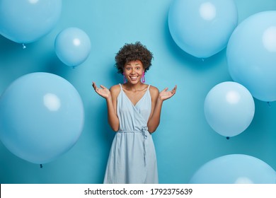 Good looking curly haired woman spreads palms, smiles sincerely, enjoys summer party, wears blue dress, stands against festive air balloons, has happy mood, isolated. Feminity, style, fashion concept