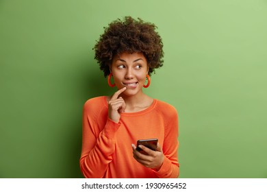 Good looking dark skinned young woman with curly hair holds modern smartphone uses new application dressed in casual orange sweater isolated over green background thinks about something pleasant
