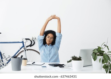 Good Looking Dark-skinned Woman Employee In Stylish Glasses Stretching Her Arms, Keeping Eyes Closed, Sitting At Modern White Office Desk With Papers And Electronic Gadgets At The End Of Working Day