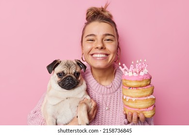 Good looking European young woman with hair bun holds pug dog and delicious doughnuts celebrates birthday has upbeat mood wears knitted jumper isolated over pink background. Holidays concept